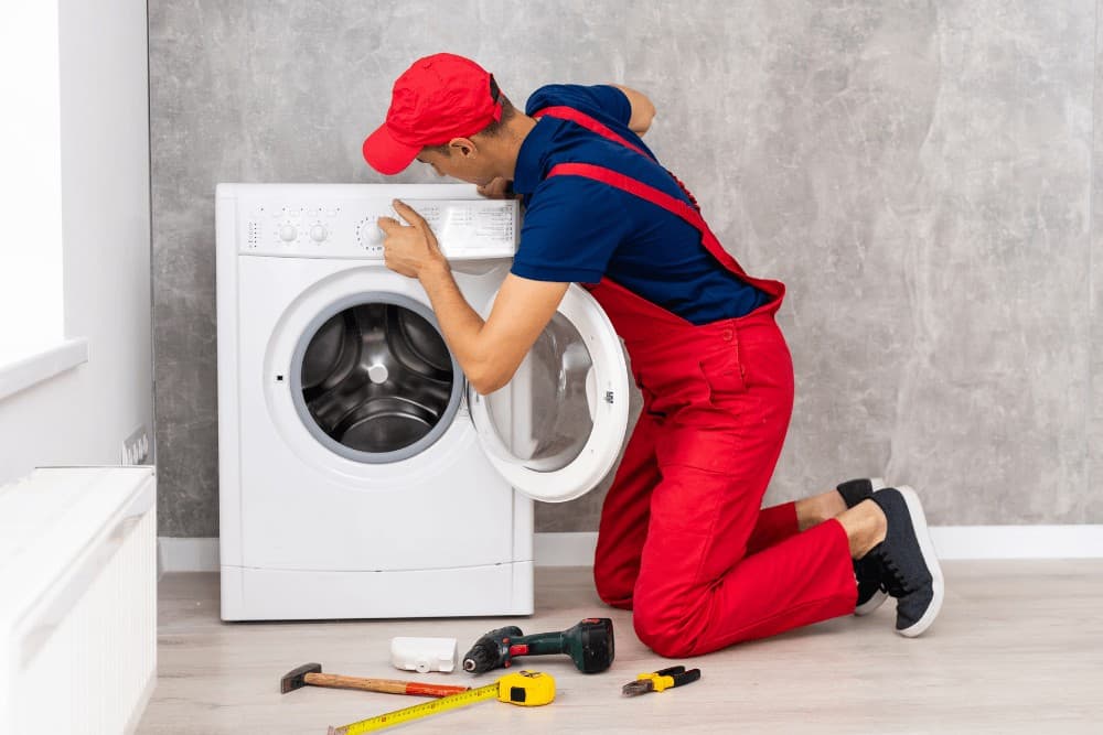 Technician in a red cap and overalls kneeling beside an open front-loading washing machine, with hand tools scattered on the floor during a repair