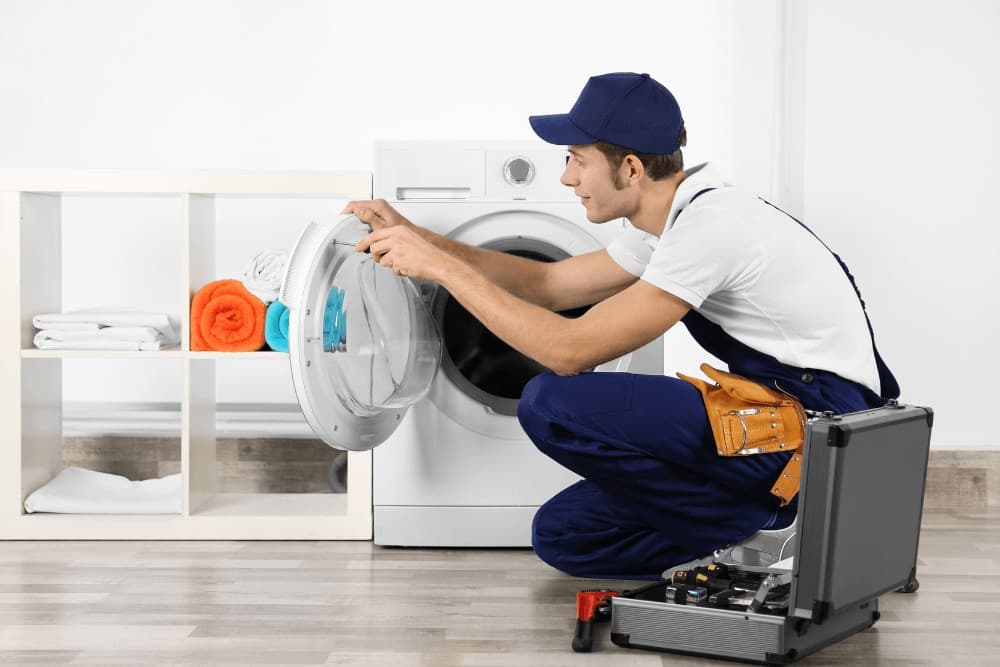 Technician in a blue cap kneeling in a utility closet, repairing a washing machine with an open toolbox on the floor beside him