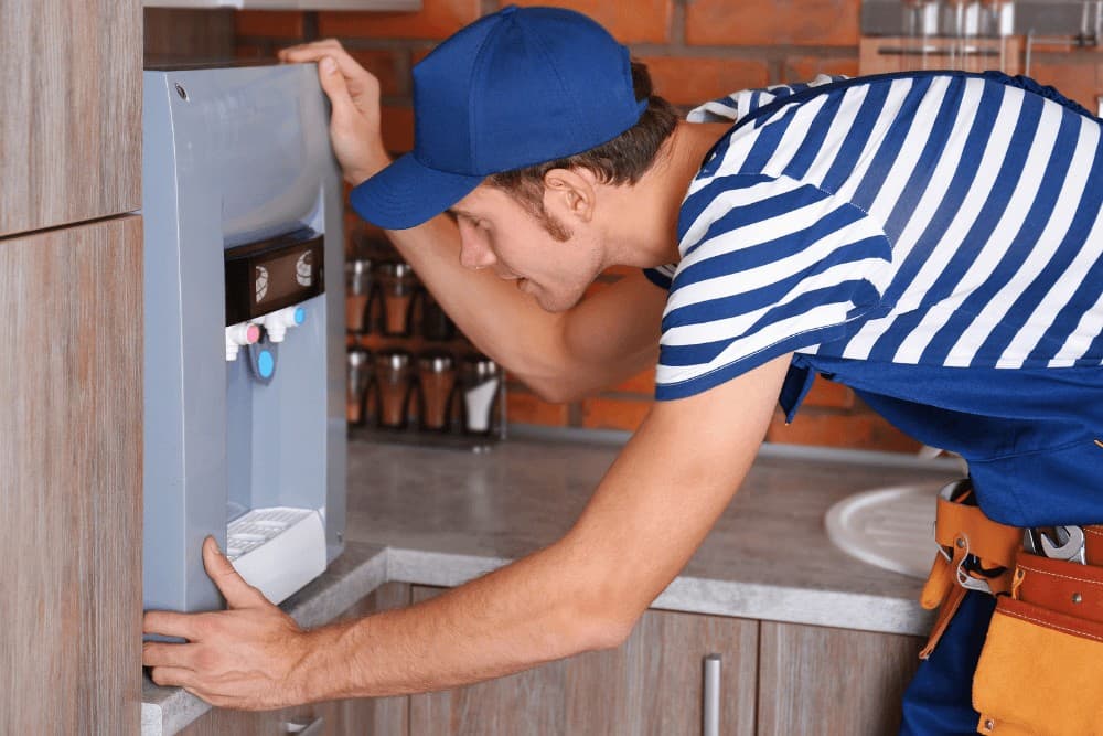 One Click Technicians technician in a striped shirt and cap leaning into a wall-mounted water dispenser unit, working on its internal tap and cooling assembly in a Abu Dhabi kitchen
