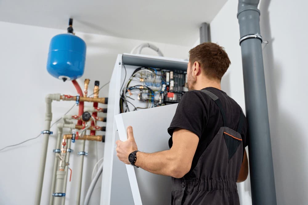 Technician checking the wiring and control panel of a wall-mounted electric water heater, with the pressure expansion vessel and copper pipework visible beside the unit in a utility room